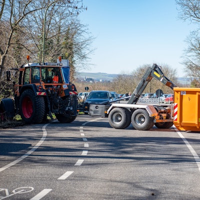 Ein Traktor mit Anhänger und ein oranger Container blockieren eine breite Straße.