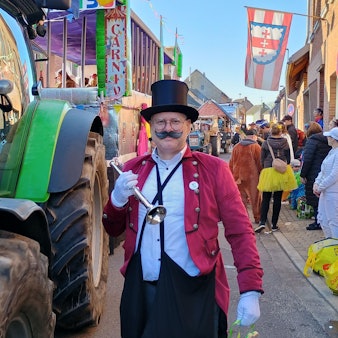 Der Mann trägt einen Zylinder, eine rote Veste und ein kleines Blechblasinstrument. Er posiert neben einem Traktor.