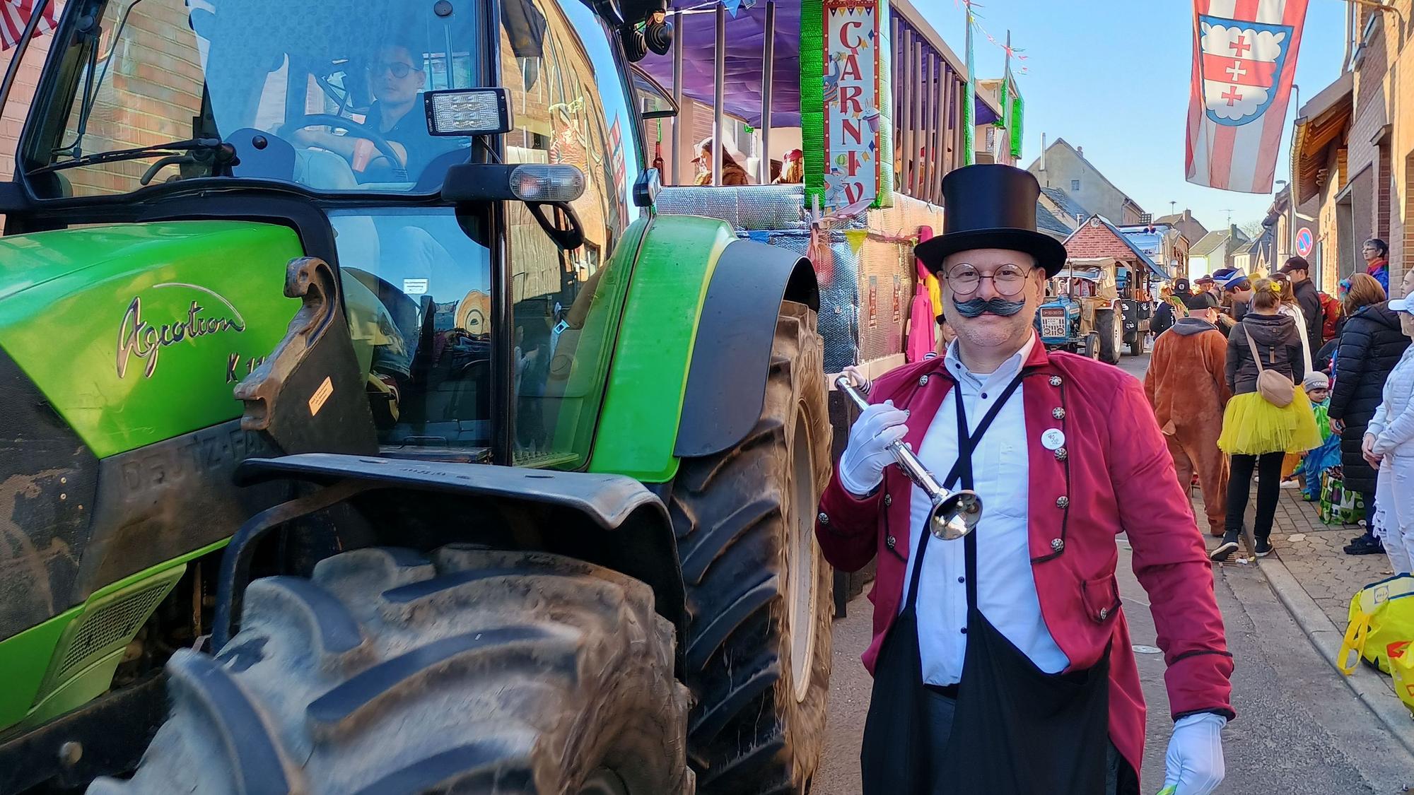 Der Mann trägt einen Zylinder, eine rote Veste und ein kleines Blechblasinstrument. Er posiert neben einem Traktor.