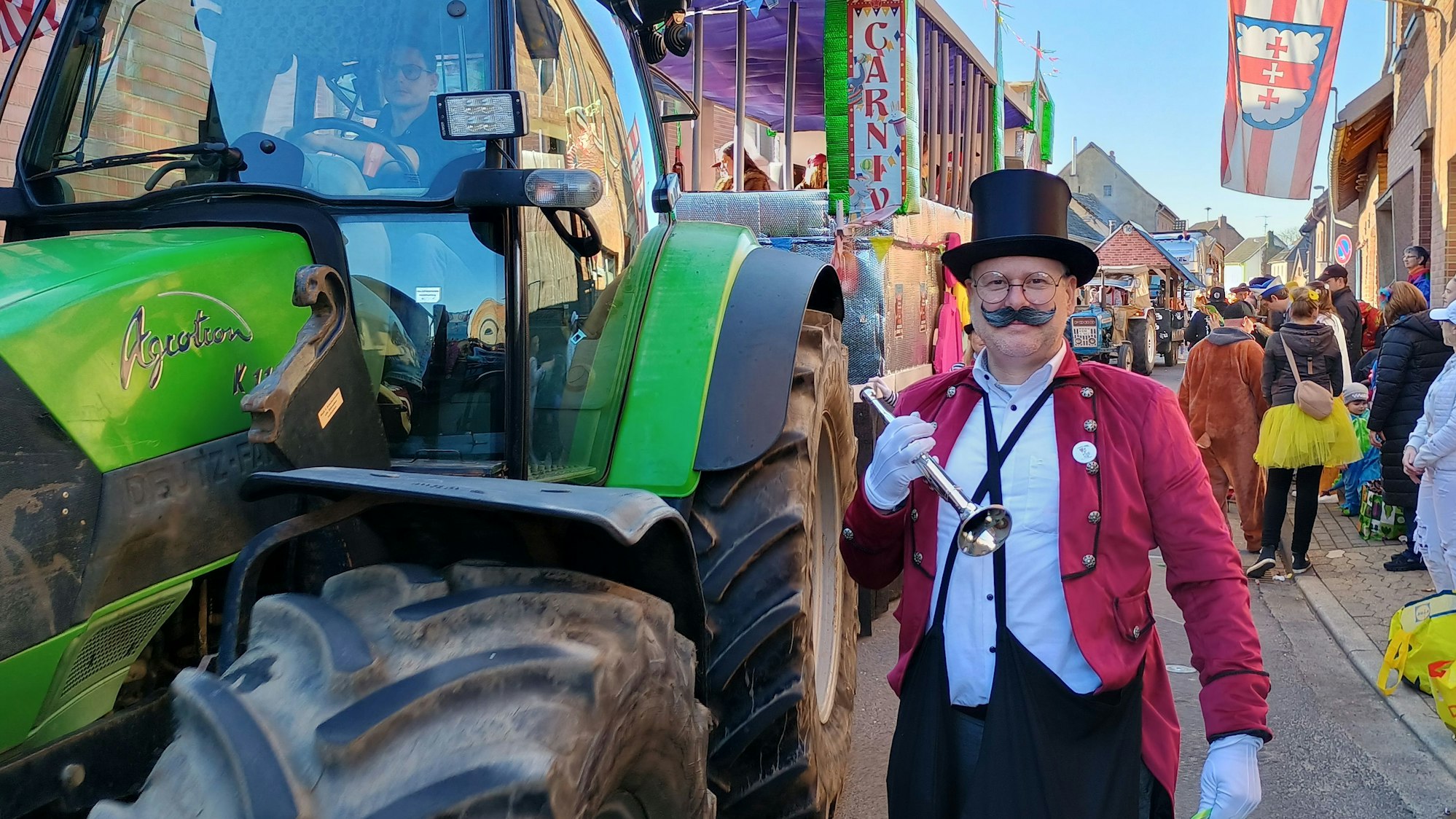 Der Mann trägt einen Zylinder, eine rote Veste und ein kleines Blechblasinstrument. Er posiert neben einem Traktor.
