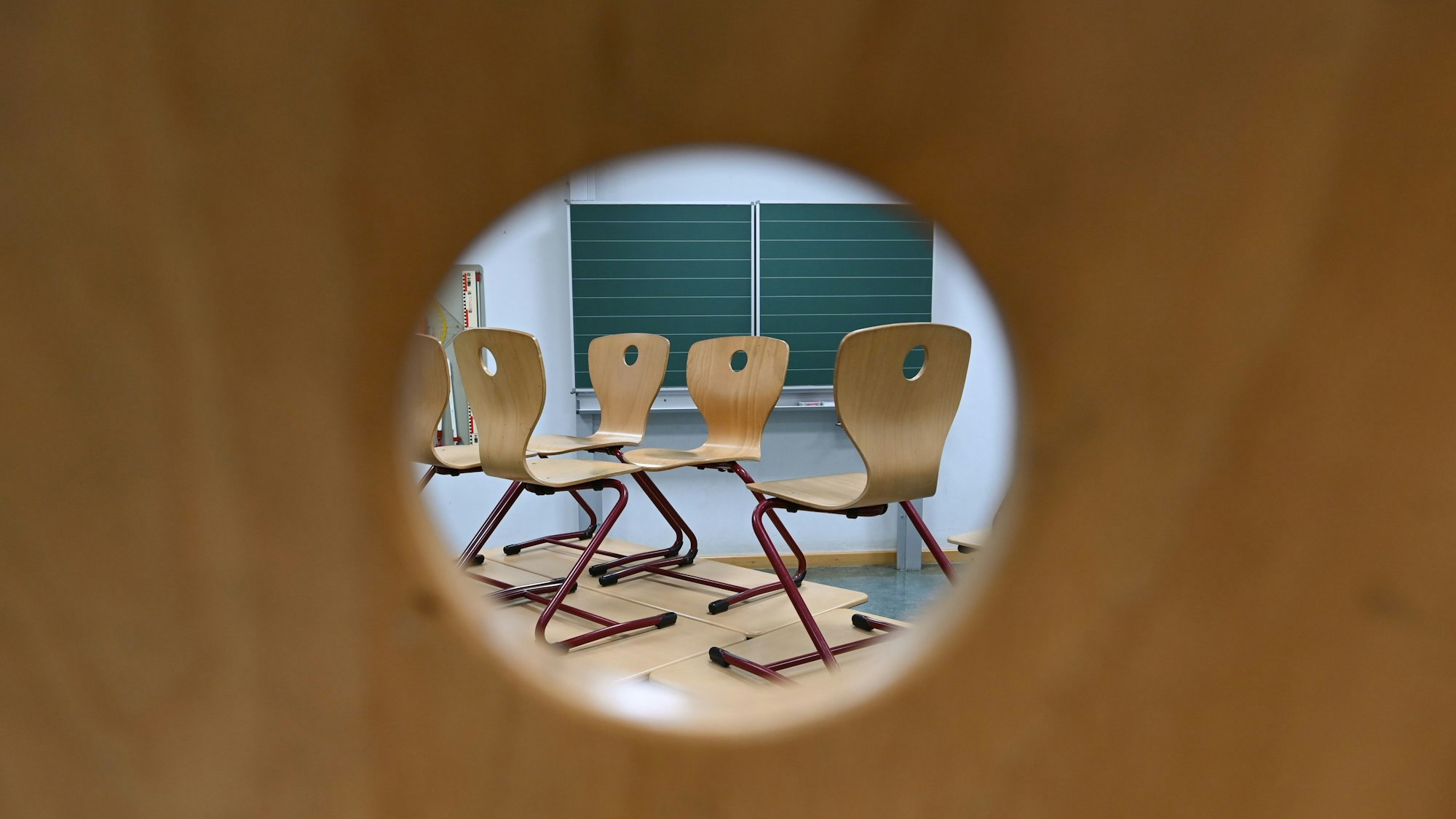Durch das Loch in einer Stuhllehne sind in einem Klassenzimmer Stühle zu sehen, die auf Tischen stehen.