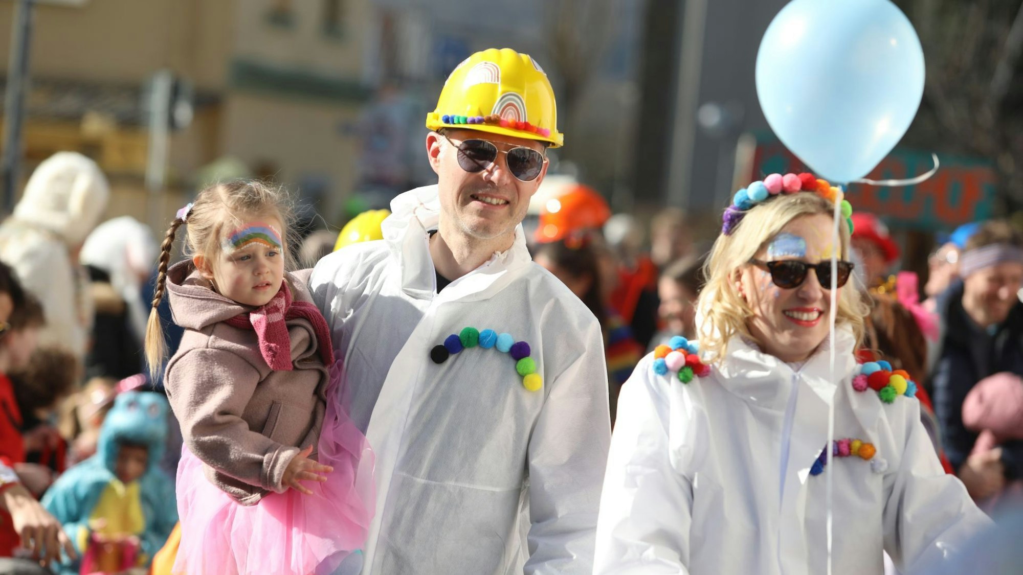 Ein Vater, der einen gelben Bauhelm und einen weißen Overall trägt, zieht mit seiner Tochter auf dem Arm durch den Ort.