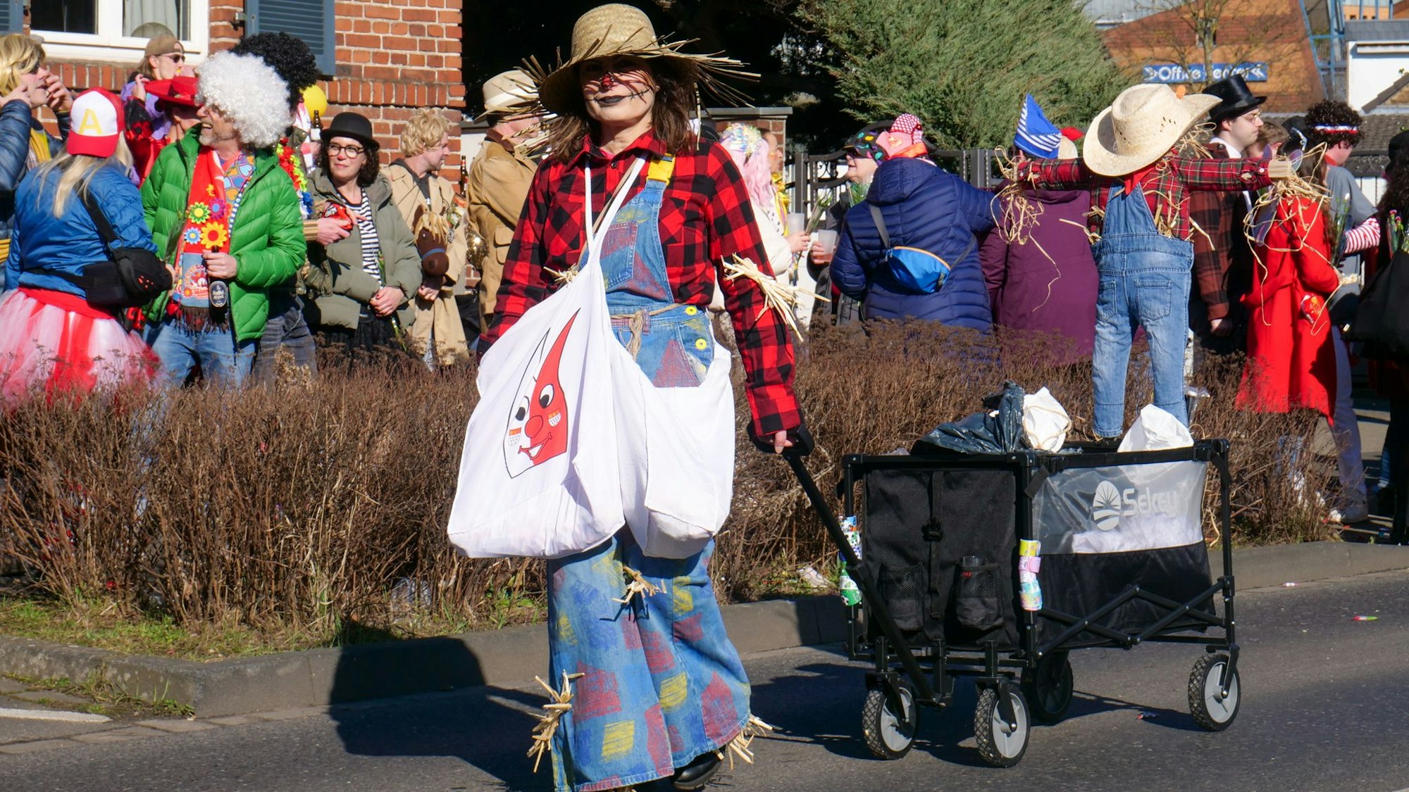 Das Bild zeigt eine Frau, die auf dem Zugweg einen Bollerwagen schiebt und ein Kostüm als Vogelscheuche trägt.