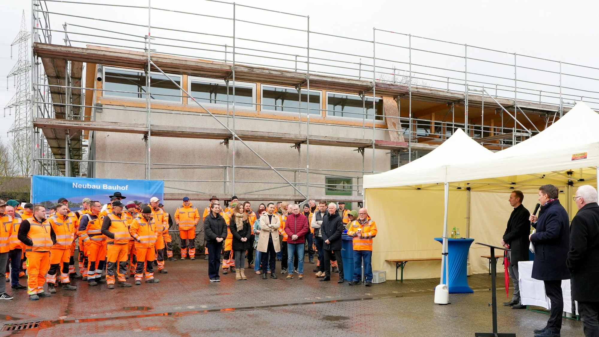 Ein eingerüstetes Gebäude, davor stehen zahlreiche Personen in orangefarbenen Jacken.