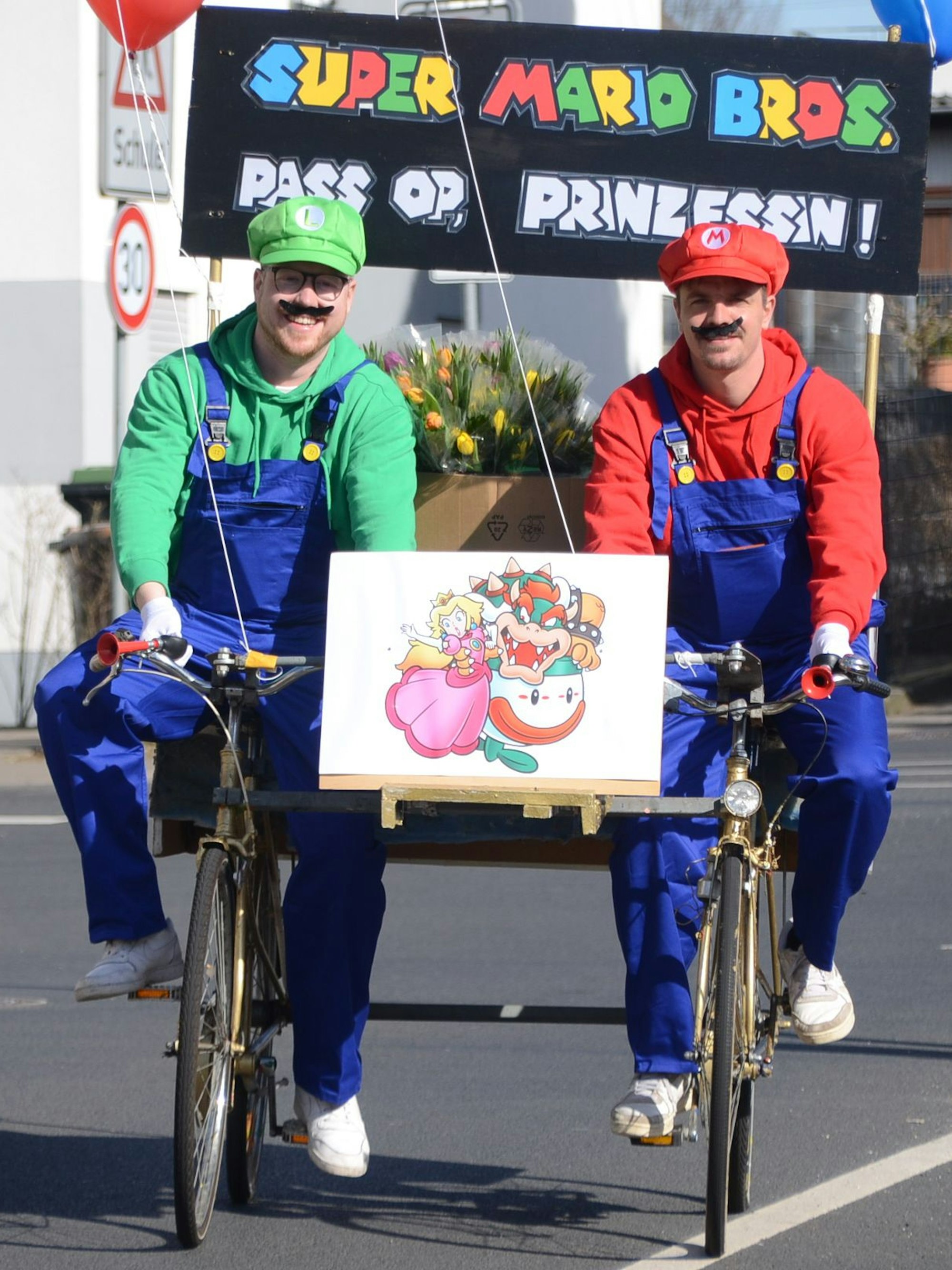 Zwei Männer in Kostümen aus dem Computerspiel Super Mario fahren auf zwei aneinander geschweißten Fahrrädern.