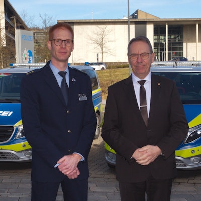 Zwei Männer stehen vor einer Polizeiwache in Gummersbach. Im Hintergrund sind zwei Polizeiautos zu sehen. Einer der Männer trägt eine Polizeiuniform.