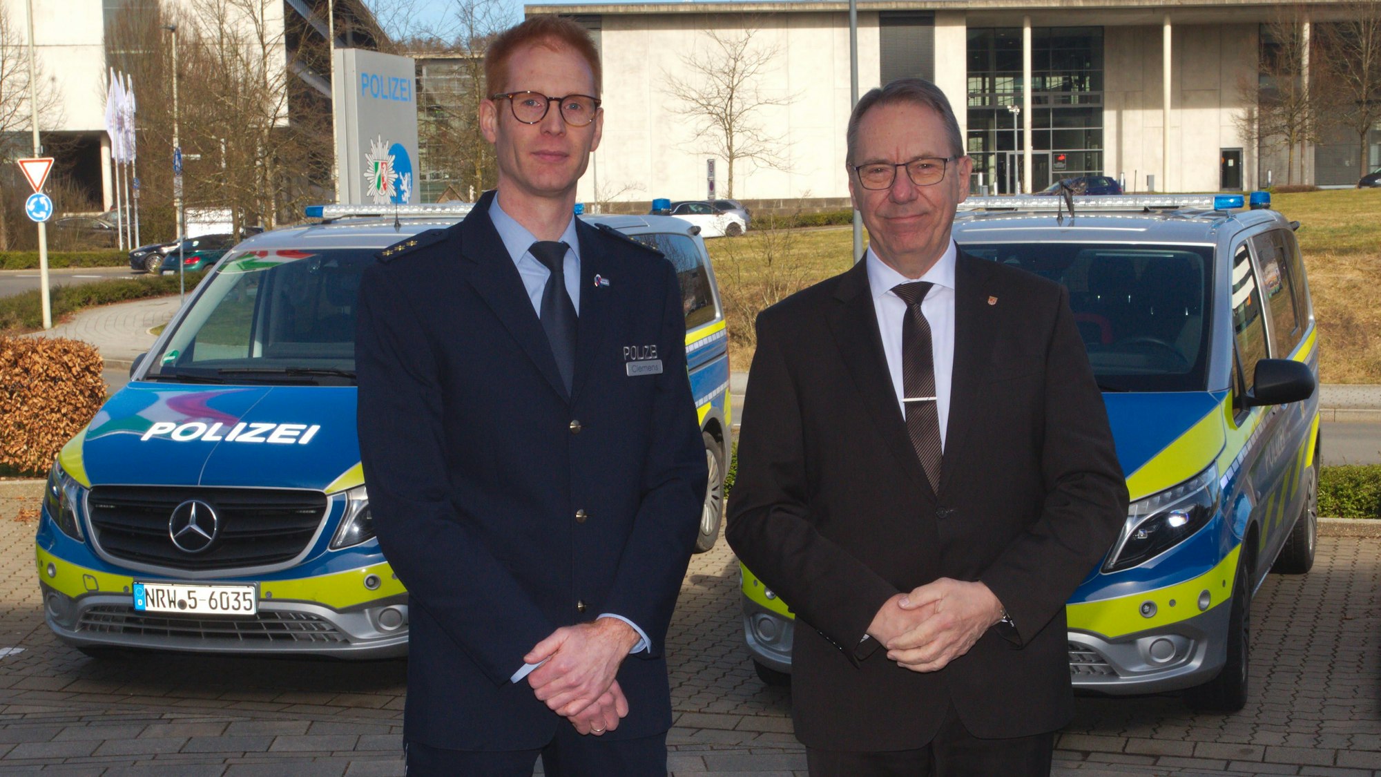 Zwei Männer stehen vor einer Polizeiwache in Gummersbach. Im Hintergrund sind zwei Polizeiautos zu sehen. Einer der Männer trägt eine Polizeiuniform.