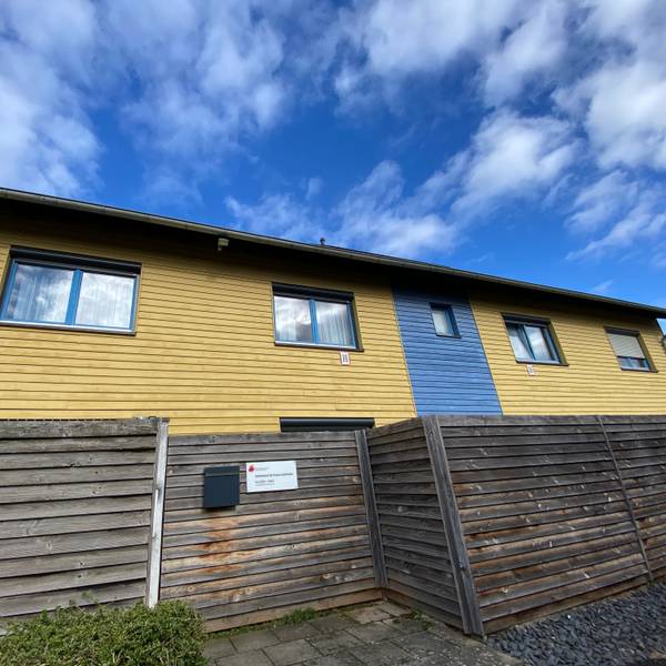Das Gebäude in den Farben Gelb und Blau besteht aus Holz, eine Holzwand ist davor angebracht.