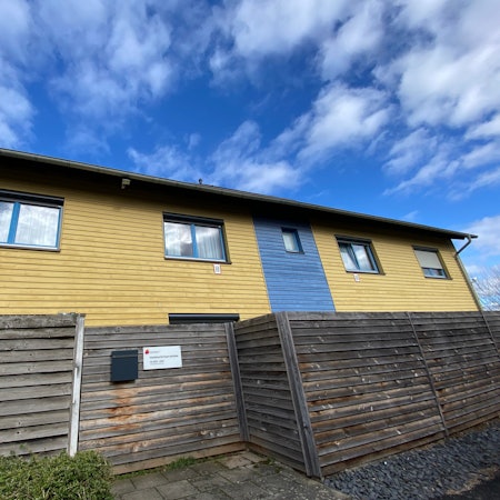 Das Gebäude in den Farben Gelb und Blau besteht aus Holz, eine Holzwand ist davor angebracht.
