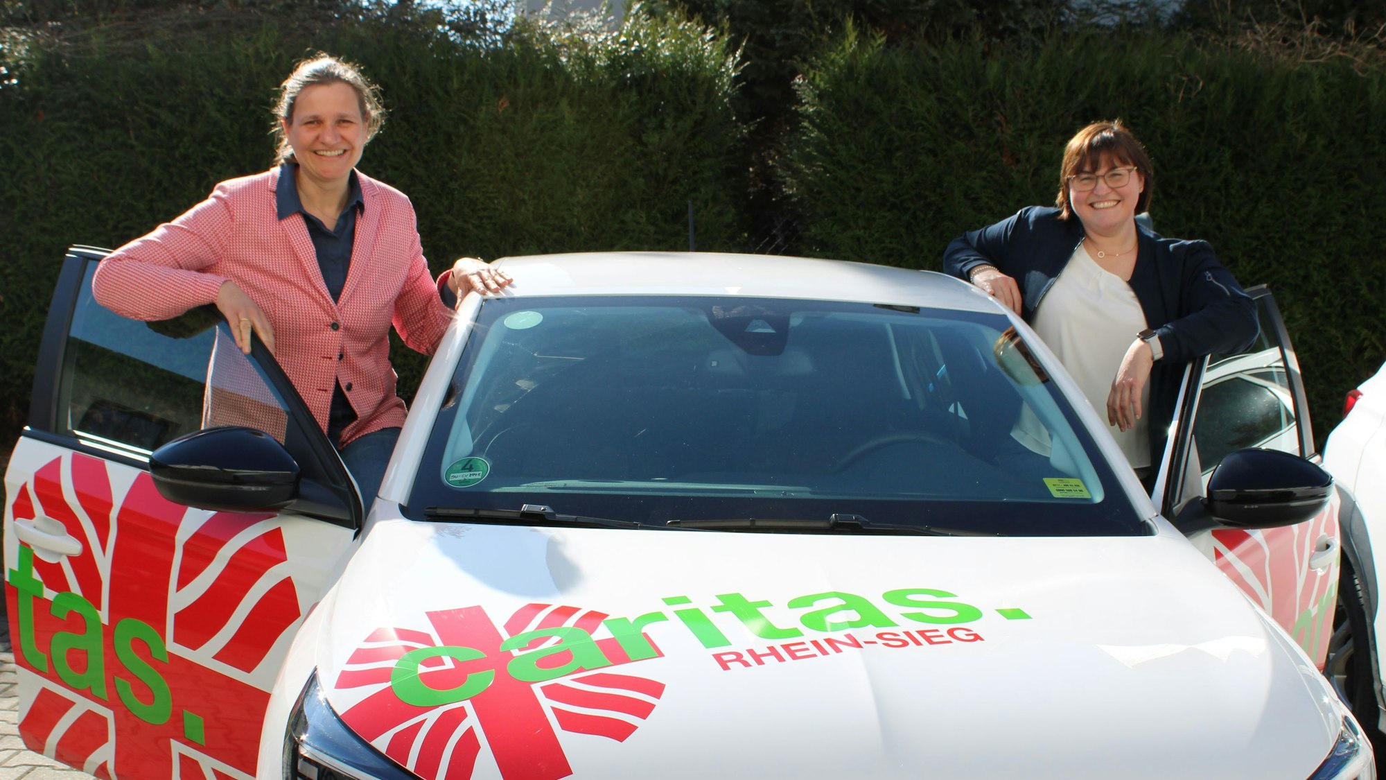 Zwei Frauen stehen an beiden vorderen Türen eines Autos, als wollten sie gerade einsteigen - auf dem Wagen steht „Caritas Rhein-Sieg“.