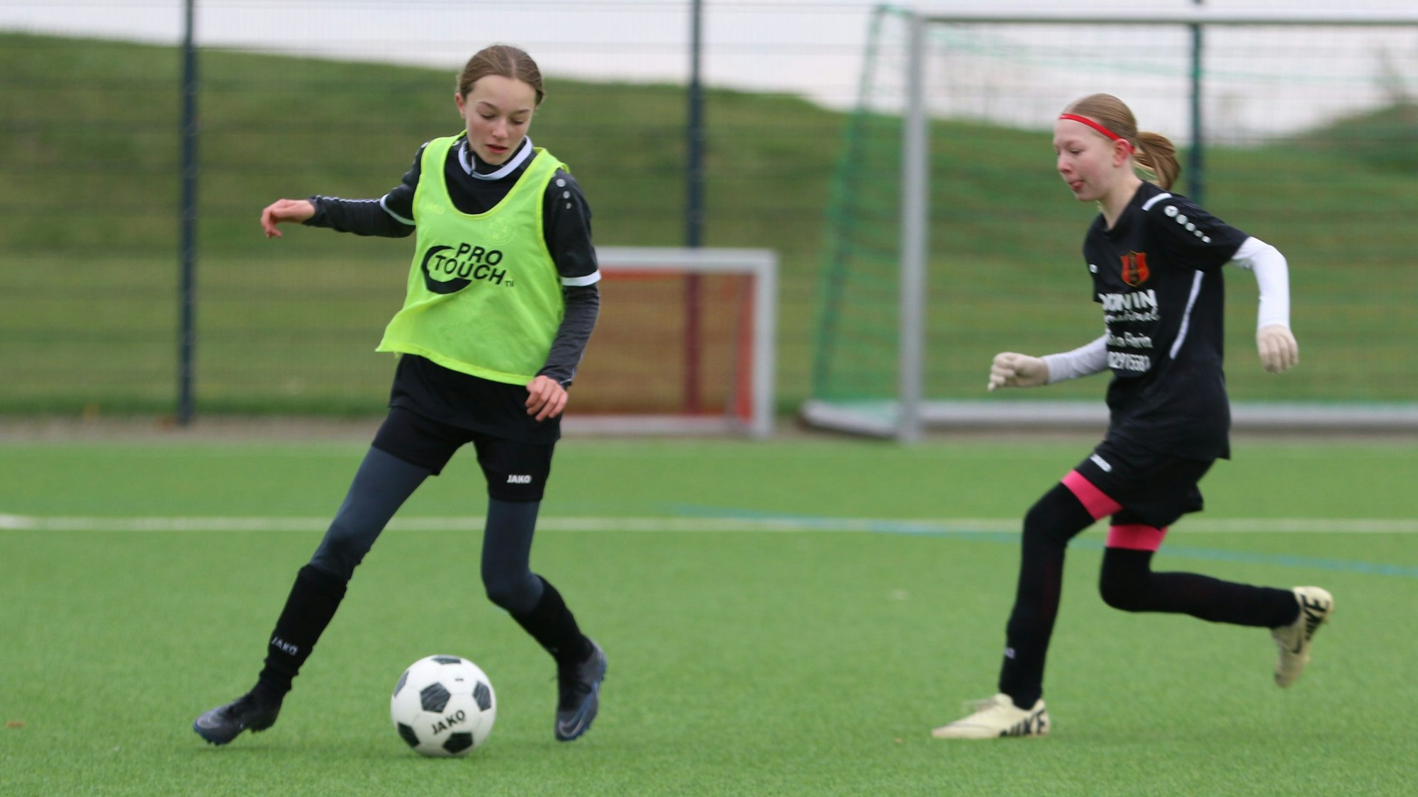 Eine Fußballerin in einem grünen Leibchen führt den Ball. Eine Gegenspielerin mit rotem Stirnband nähert sich ihr.