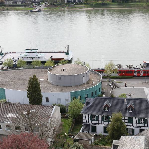 Ein ovales Gebäude mit grüner Fassade, dahinter der Rhein und die Fähre Königswinter.