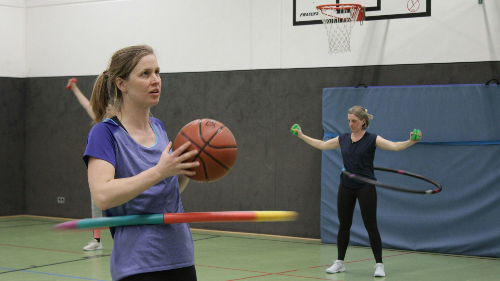 Eine Frau wirft einen Basketball und schaut nach oben, während sie einen Hula Hoop Reifen Kreisen lässt. Im Hintergrund hebt eine andere Frau Gewichte.