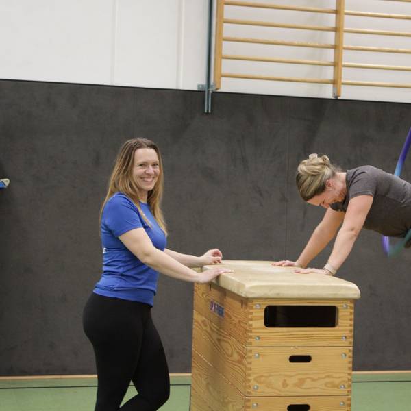Eine Frau ist auf zwei Kasten-Turngeräte gestützt und lässt in Plank-Position einen Reifen Kreisen. Eine andere Frau steht lächelnd neben ihr.