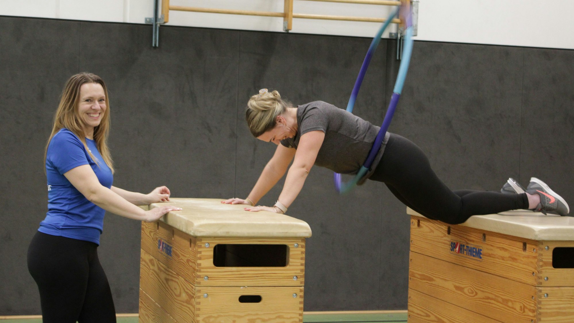 Eine Frau ist auf zwei Kasten-Turngeräte gestützt und lässt in Plank-Position einen Reifen Kreisen. Eine andere Frau steht lächelnd neben ihr.