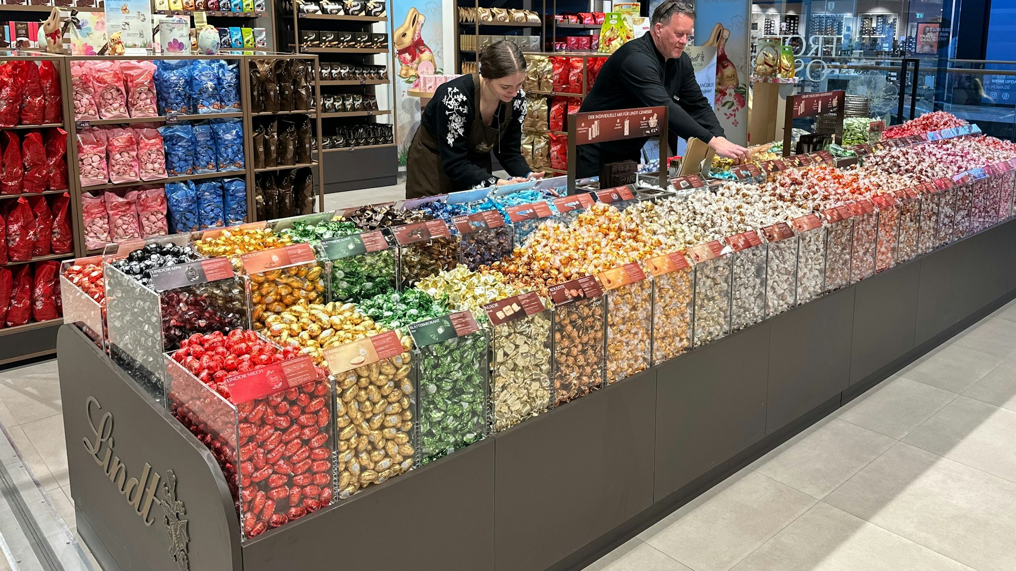 Blick in ein Süßwarenoutlet. In Kunststoffschütten sind große Mengen bunt verpackter Schokoladenkugeln zu sehen.