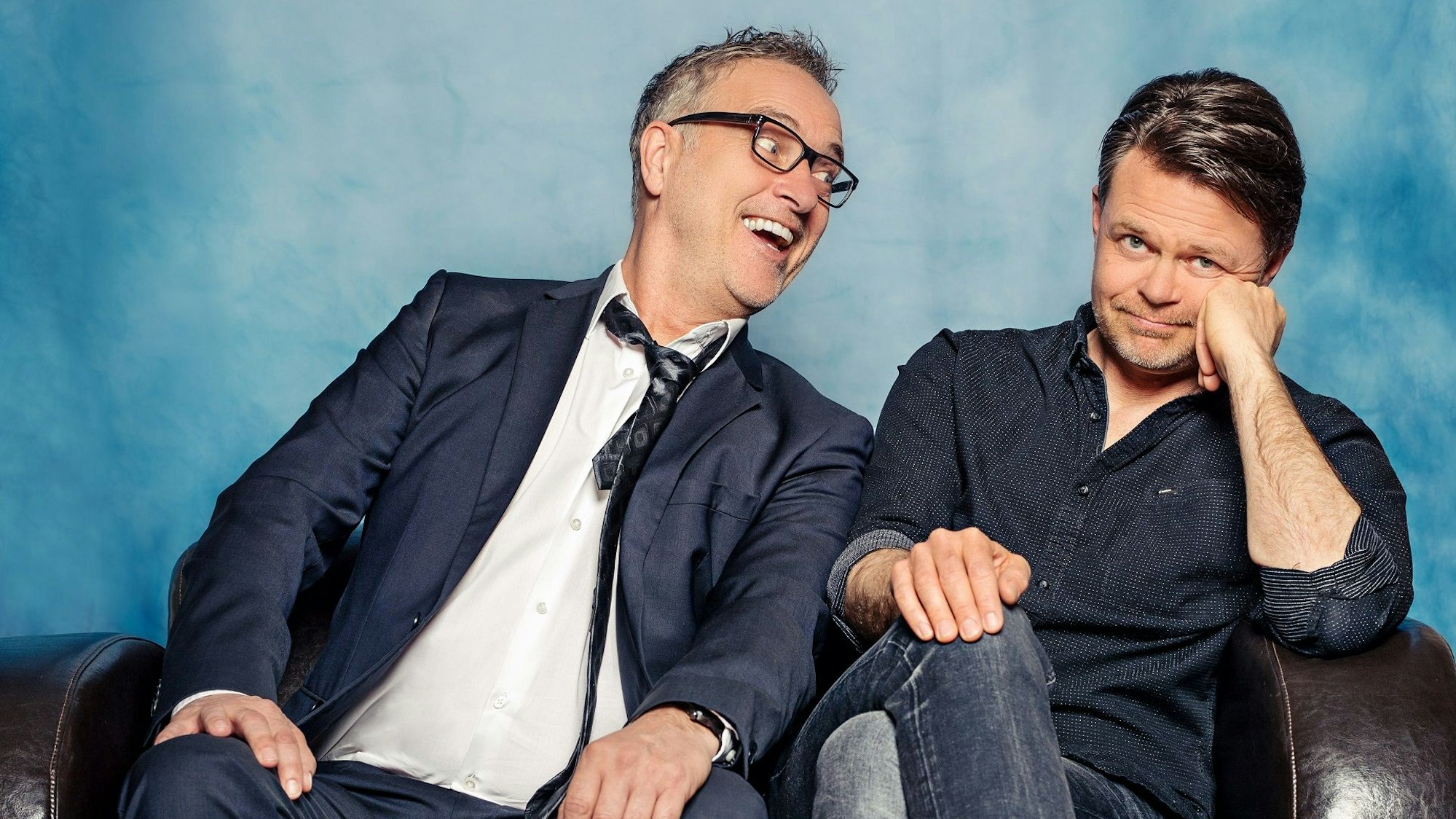 Zwei Herren sitzen auf einem Sofa, der linke lacht, der rechte guckt zweifelnd.