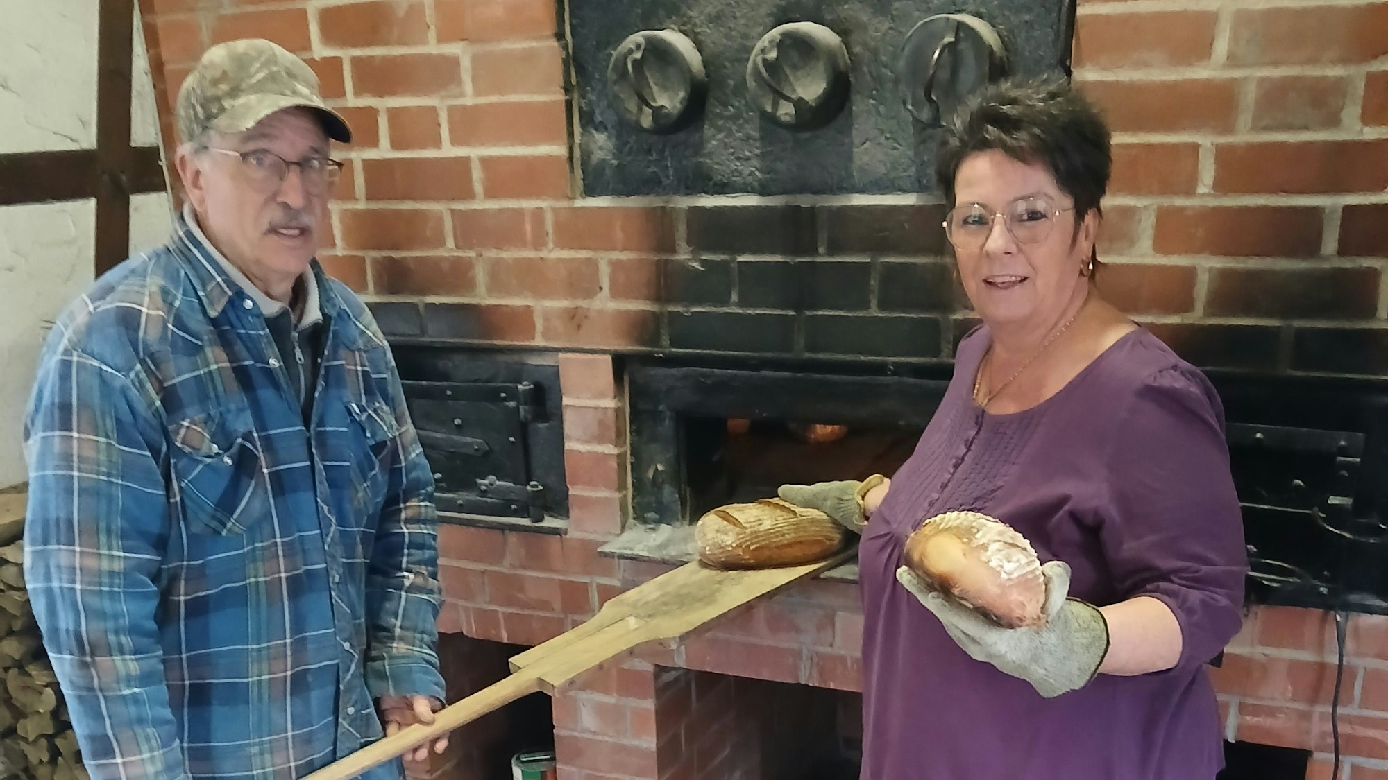 Ein Mann und eine Frau stehen an einem Steinbackofen. Er zieht auf einem Schieber ein Brot aus dem Ofen, sie hält eines in der Hand.
