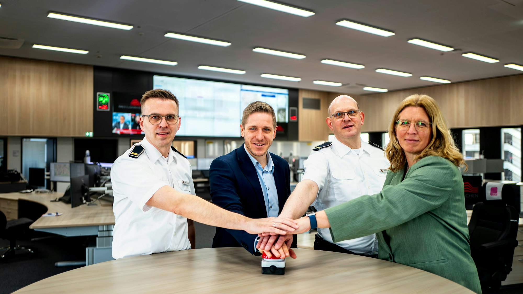 Drei Männer und eine Frau drücken auf einen roten Buzzer, im Hintergrund sind die Arbeitsplätze der Disponenten und die großen Bildschirme an der Wand zu erkennen.