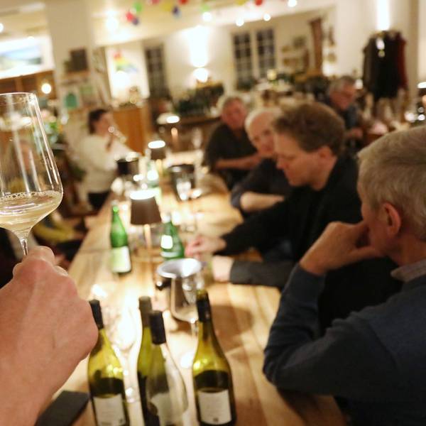 Ein Weinglas wird in die Höhe gehoben, um zu zeigen, dass der trübe Wein noch jung ist. Auf dem Tisch stehen weitere Flaschen.