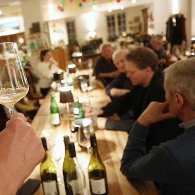 Ein Weinglas wird in die Höhe gehoben, um zu zeigen, dass der trübe Wein noch jung ist. Auf dem Tisch stehen weitere Flaschen.