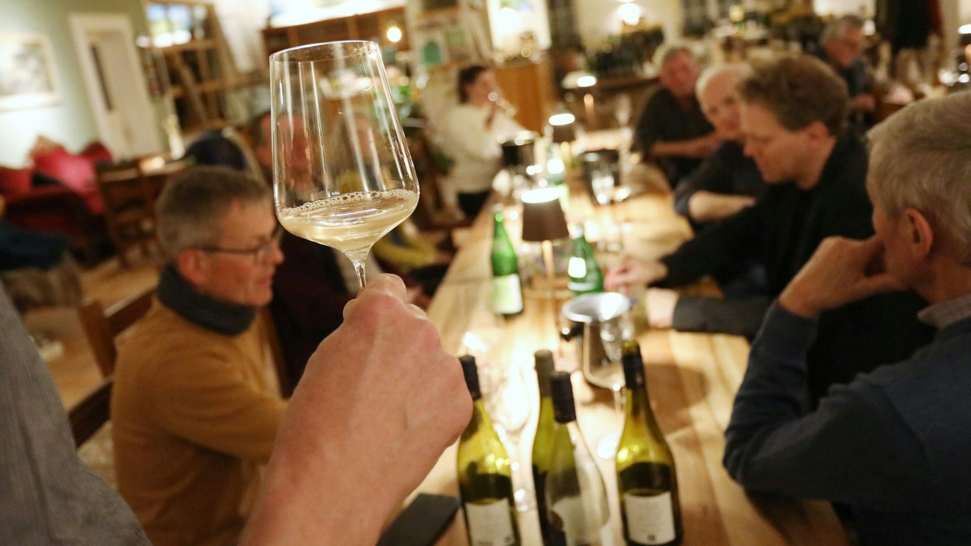 Ein Weinglas wird in die Höhe gehoben, um zu zeigen, dass der trübe Wein noch jung ist. Auf dem Tisch stehen weitere Flaschen.