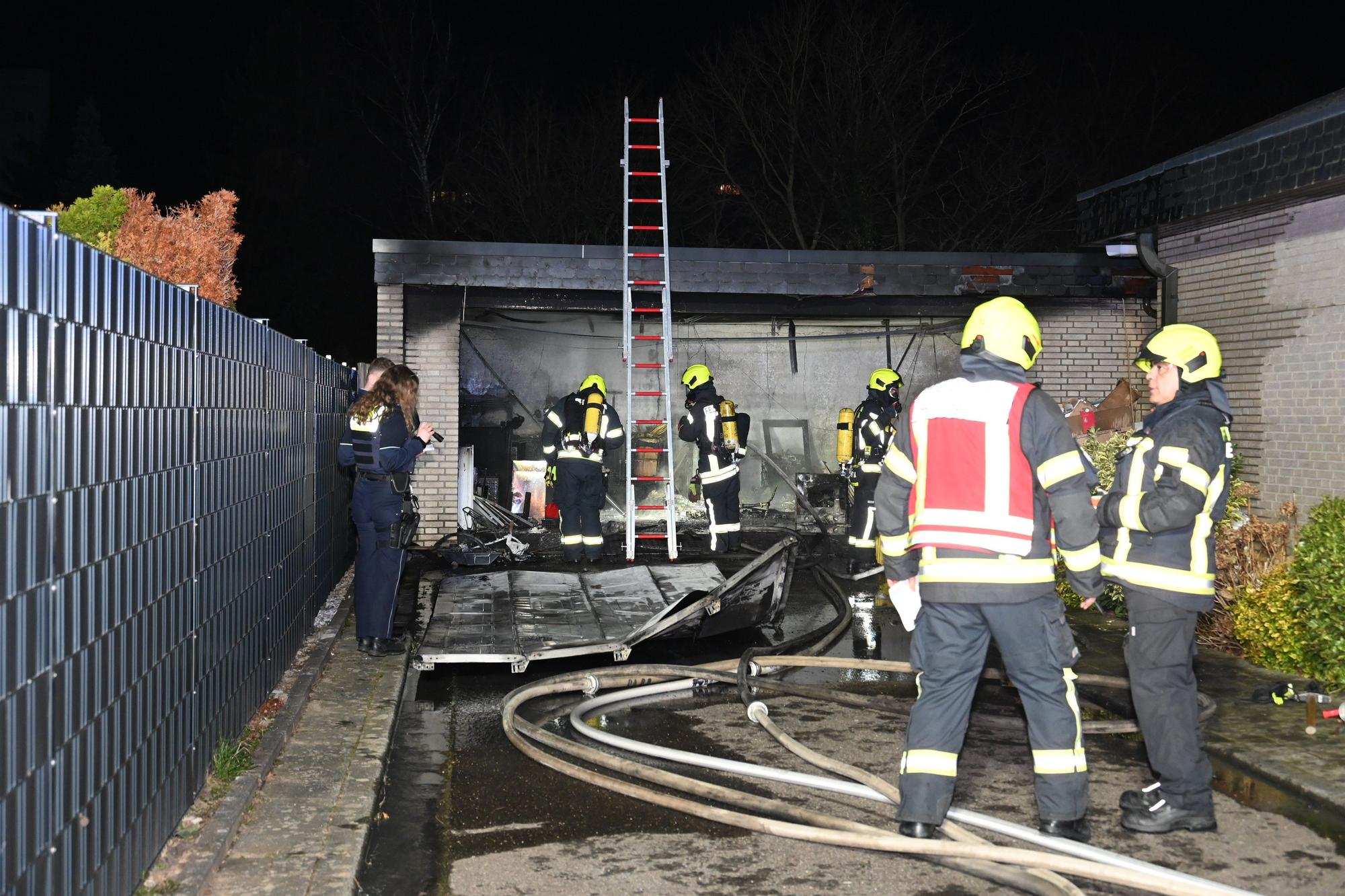 Feuerwehrleute stehen vor einer ausgebrannten Garage. Schläuche liegen auf dem Boden und das zerstörte Tor liegt vor der Garage.