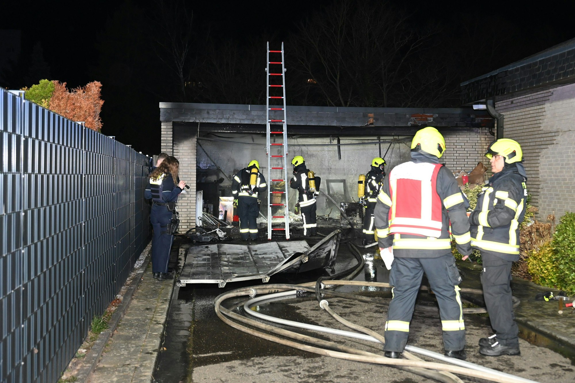 Feuerwehrleute stehen vor einer ausgebrannten Garage. Schläuche liegen auf dem Boden und das zerstörte Tor liegt vor der Garage.