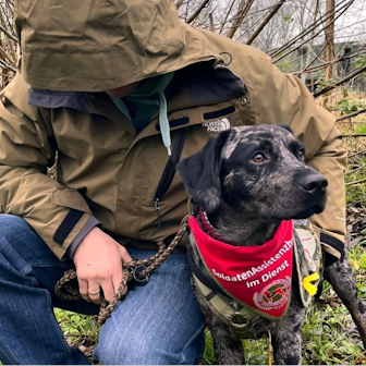 Das Foto zeigt einen Mann mit einer Kapuze, die sein Gesicht verdeckt, der den Arm um eine dunkel gefleckte Hündin gelegt hat.