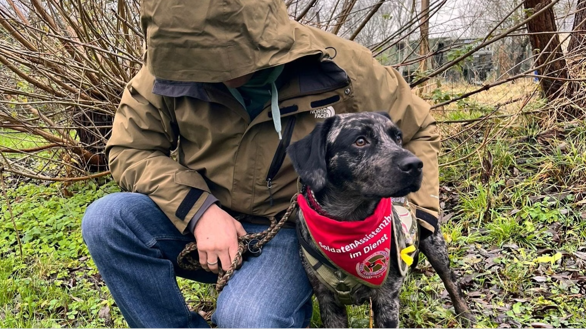 Das Foto zeigt einen Mann mit einer Kapuze, die sein Gesicht verdeckt, der den Arm um eine dunkel gefleckte Hündin gelegt hat.