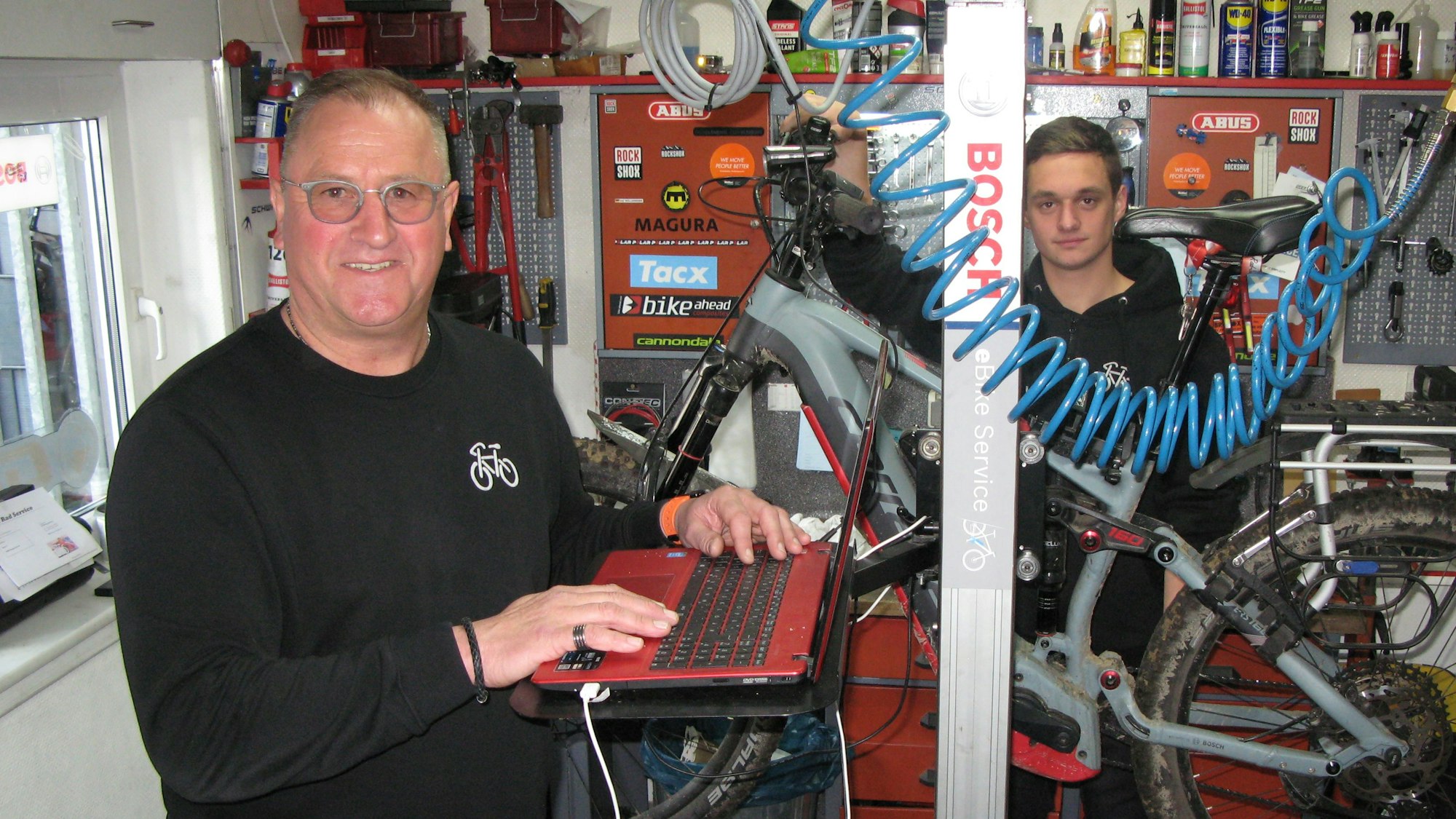 Zwei Männer stehen in einer Fahrradwerkstatt mit einem Fahrrad vor einem Regal.