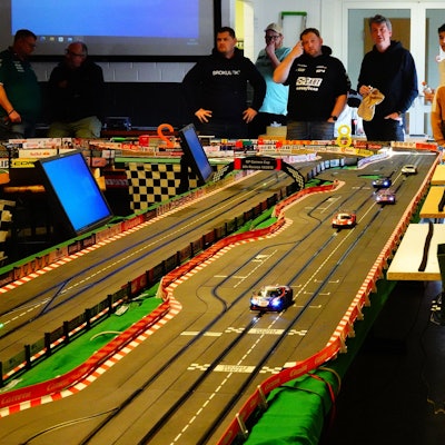 Auf dem Bild ist die Bahn zu sehen, die wie eine Rennstrecke auch zahlreiche Tafeln von mit Autos verbundenen Sponsoren zu sehen sind.  Hinter der Bahn stehen die Teilnehmer des Rennens in einer Reihe.