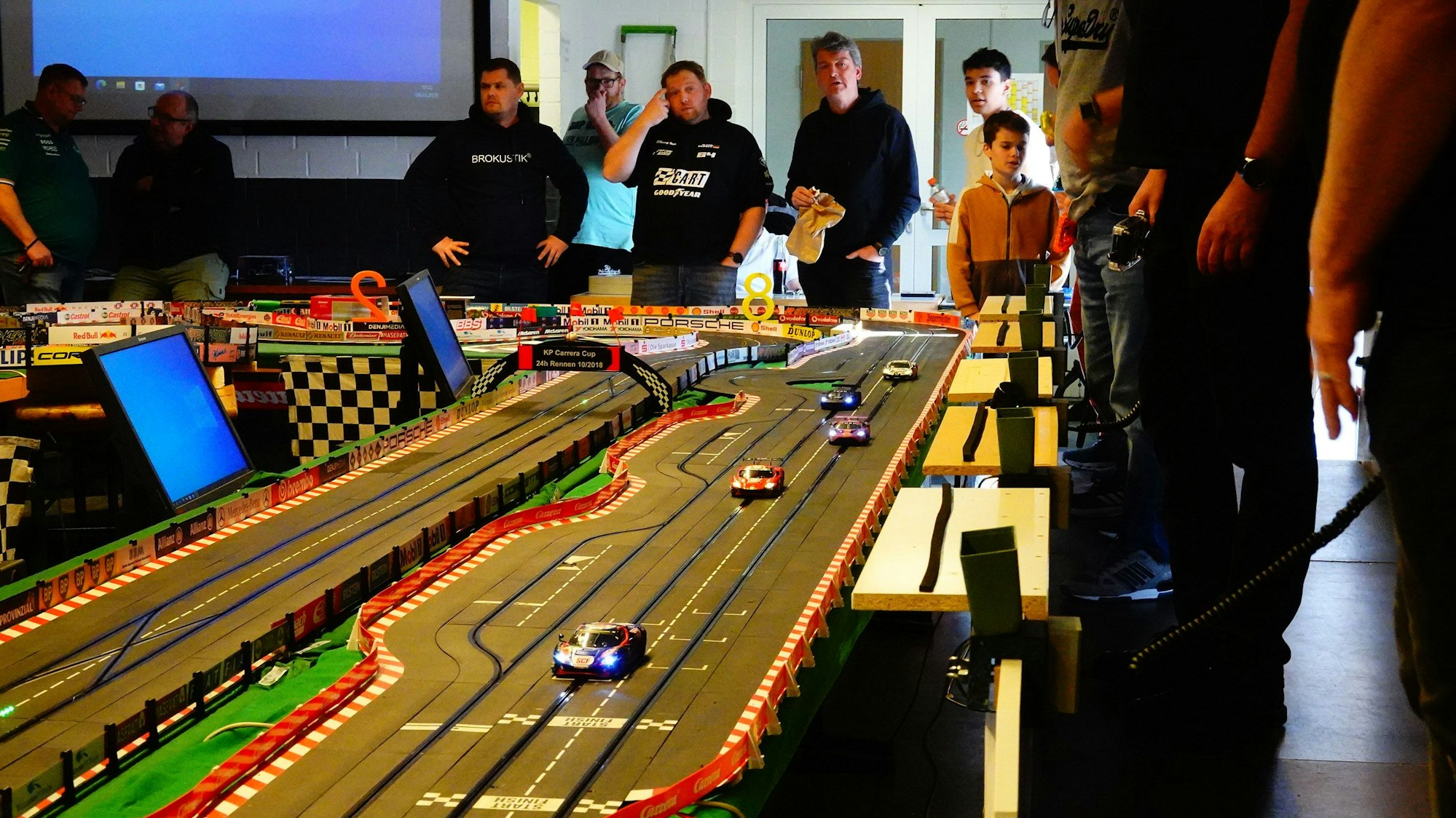 Auf dem Bild ist die Bahn zu sehen, die wie eine Rennstrecke auch zahlreiche Tafeln von mit Autos verbundenen Sponsoren zu sehen sind. Hinter der Bahn stehen die Teilnehmer des Rennens in einer Reihe.