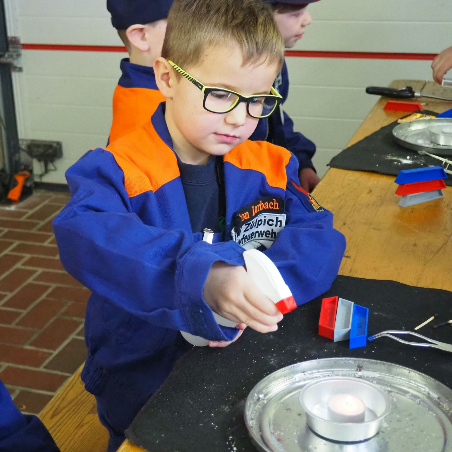 Ein Junge in orange-blauer Jacke sprüht mit einer Wassersprühflasche auf ein brennendes Teelicht.