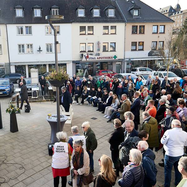 Eine Gruppe von Menschen steht auf einem Platz vor einer eingehüllten Tafel, die von sechs Metallstelen umgeben ist.