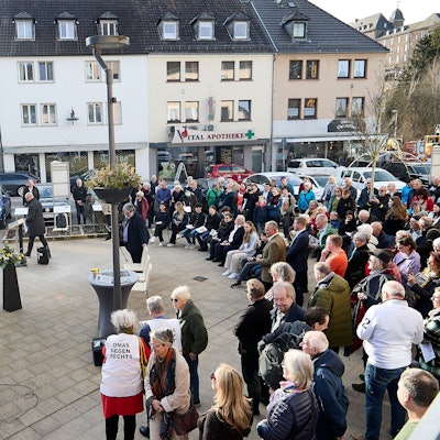 Eine Gruppe von Menschen steht auf einem Platz vor einer eingehüllten Tafel, die von sechs Metallstelen umgeben ist.