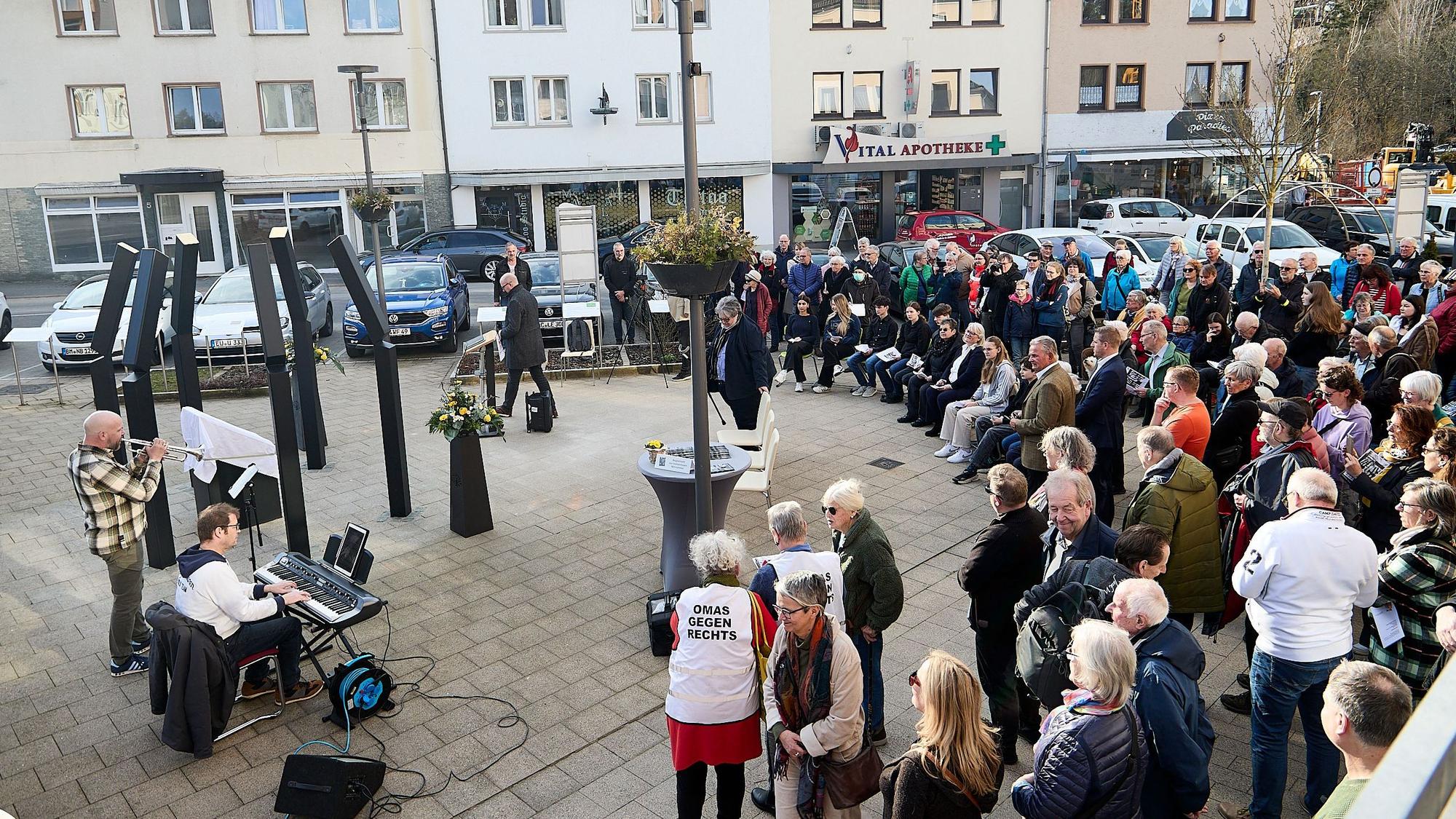 Eine Gruppe von Menschen steht auf einem Platz vor einer eingehüllten Tafel, die von sechs Metallstelen umgeben ist.