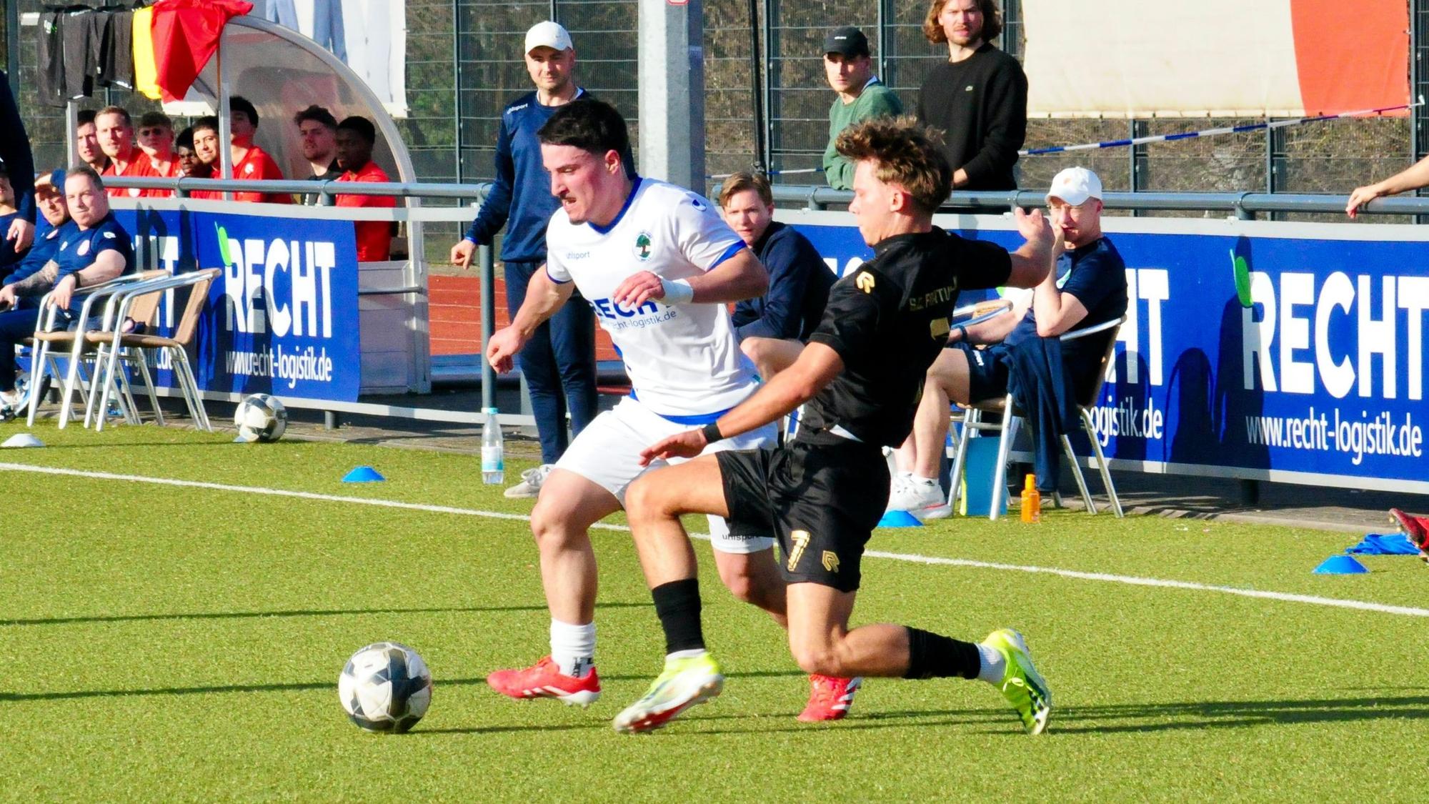 Das Foto zeigt einen Fußballer im weißen Trikot mit Ball am Fuß, der seinem Gegenspieler im schwarzen Dress bedrängt wird.