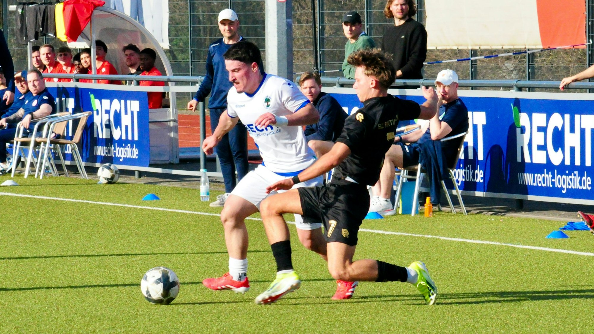 Das Foto zeigt einen Fußballer im weißen Trikot mit Ball am Fuß, der seinem Gegenspieler im schwarzen Dress bedrängt wird.