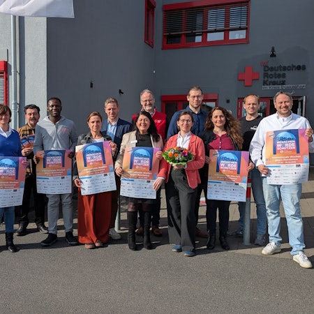 Eine Gruppe Menschen steht vor einem Gebäude des Deutschen Roten Kreuzes und hält Plakate in die Kamera.