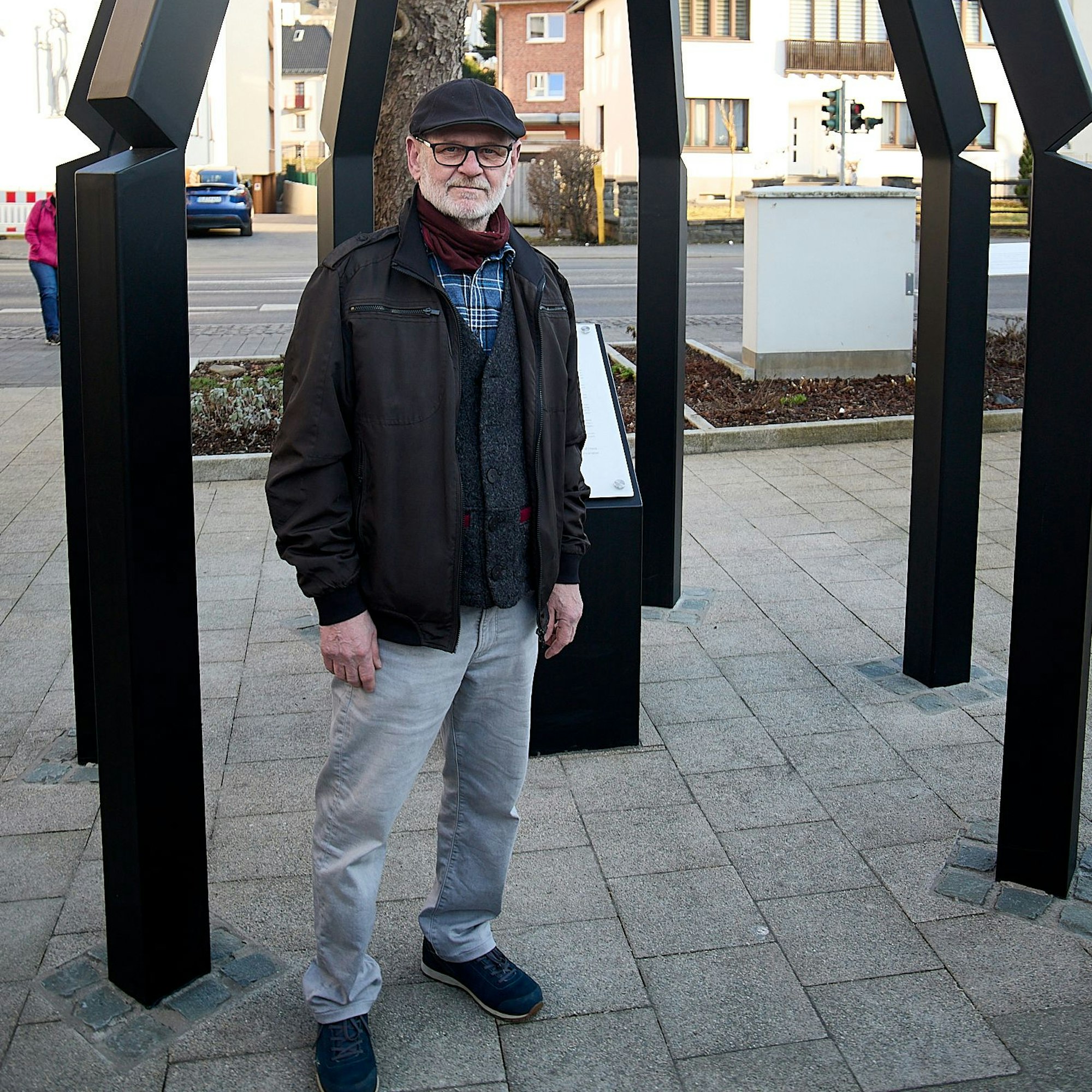 Der Schmied mit Schirmmütze steht vor der Gedenktafel, um ihn herum sind die Metallstelen zu sehen. Er trägt eine graue Hose und eine dunkle Jacke.