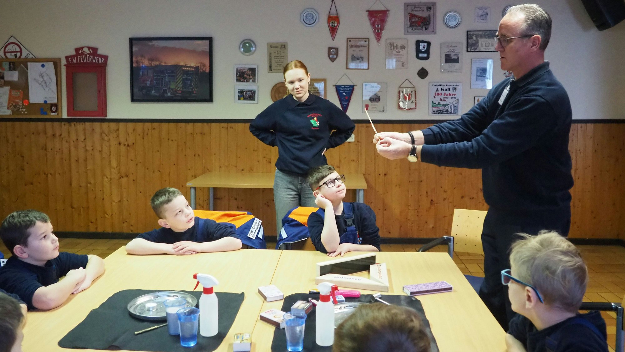 Kinder sitzen um einen Tisch, an dem ein Mann etwas mit einem Streichholz vorführt.