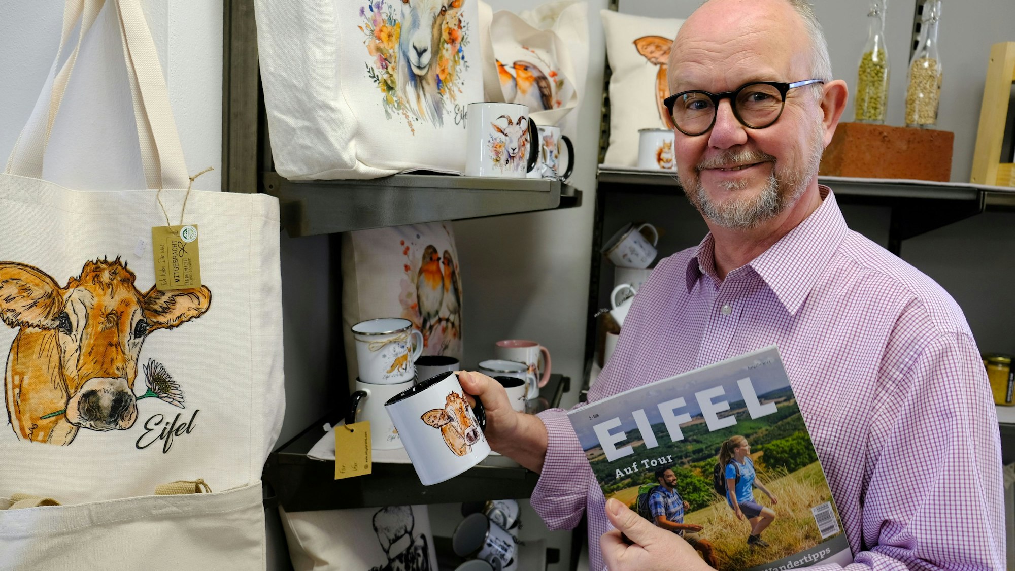 Der Schriftsteller Ralf Kramp steht vor einem Regal mit Eifel-Merch. Unter seinem linken Arm klemmt ein Tourismus-Magazin mit der Aufschrift „Eifel“.