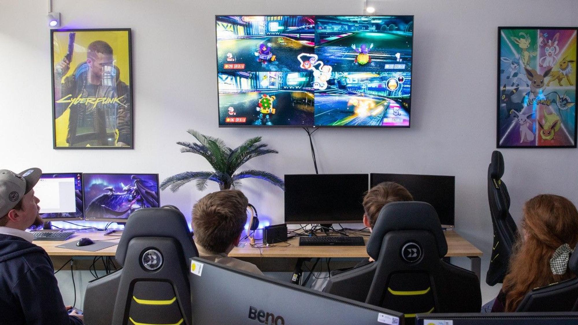 Junge Leute in speziellen Schreibtischstühlen sitzen vor einem großen Computerbildschirm, auf dem ein Spiel zu sehen ist.