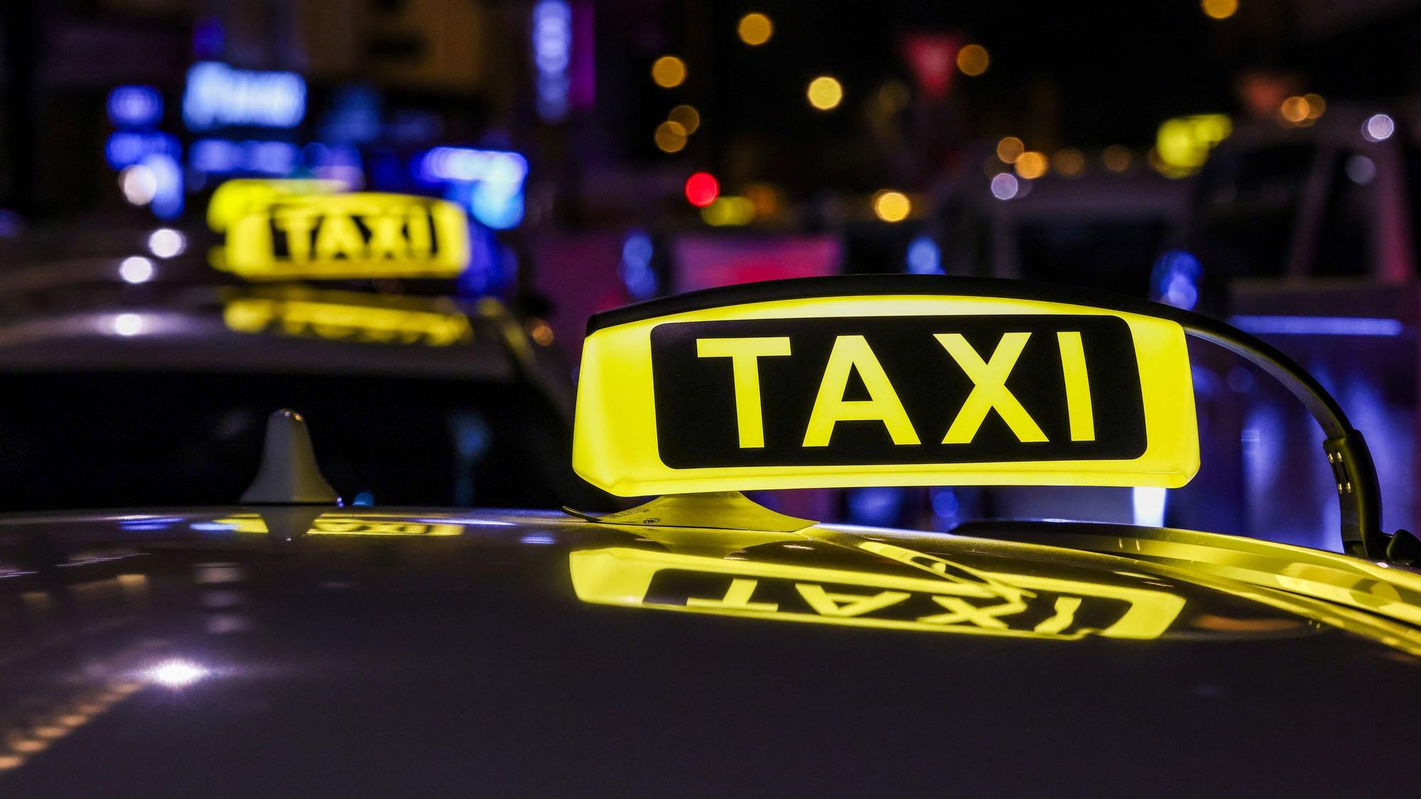 Das Bild zeigt bei Nacht ein beleuchtetes Schild mit der Aufschrift „Taxi“ auf dem Dach eines Autos.