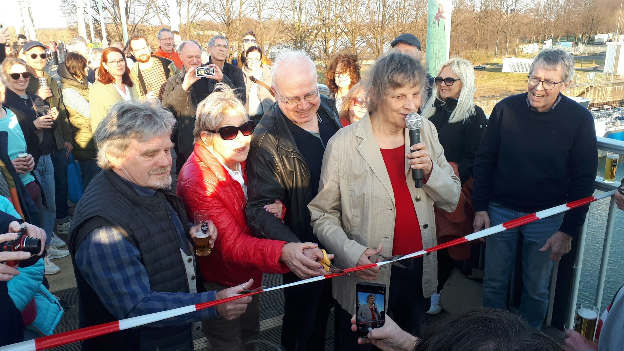 Menschen stehen auf einer Hafenbrücke. Eine Frau, die ein Mikrofon hält, schneidet ein rot-weißes Flatterband durch.