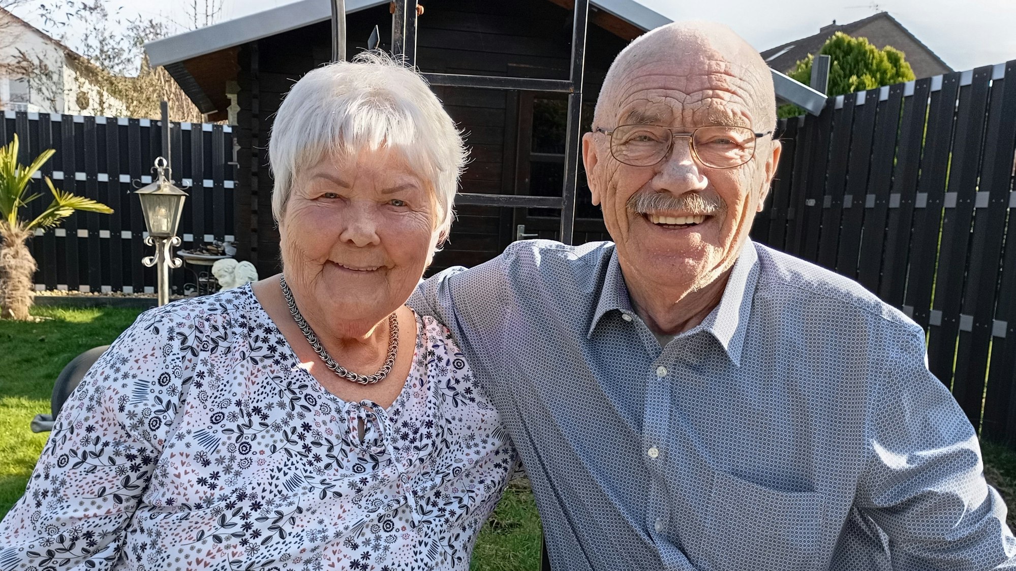 Johanna Ritz trägt eine blumige Bluse, eine Halskette und hat weiße Haare. Wilhelm Ritz trägt eine Brille und einen Schnurbart, er hat eine Glatze. Er trägt ein Hemd.