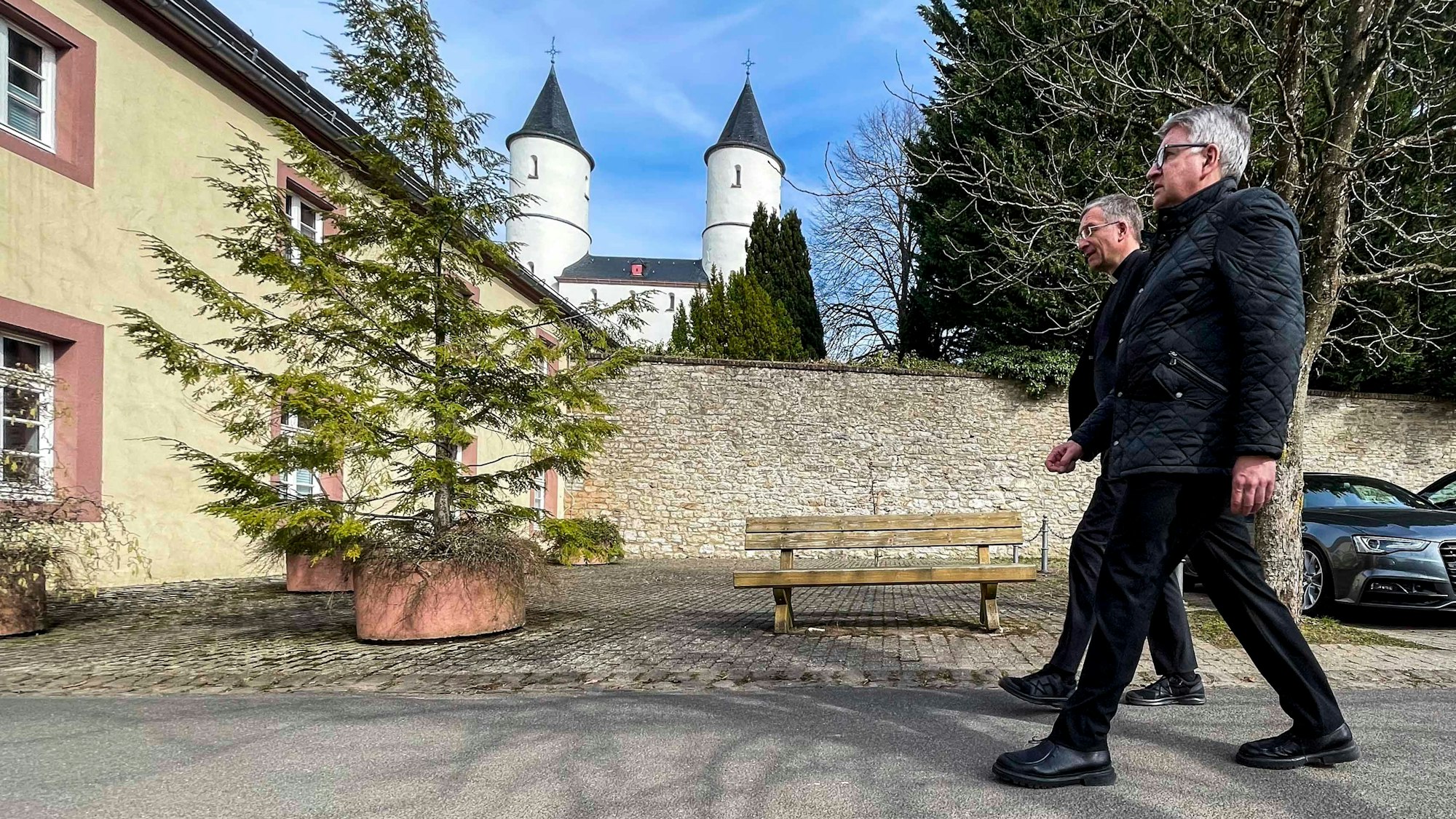 Zwei in schwarz gekleidete Männer gehen in Kall-Steinfeld einen Weg entlang. Im Hintergrund sind die Türme der Basilika zu sehen.