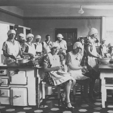 Schwarzweiß-Foto von jungen Frauen mit Haube und Schürze in einer Küche.