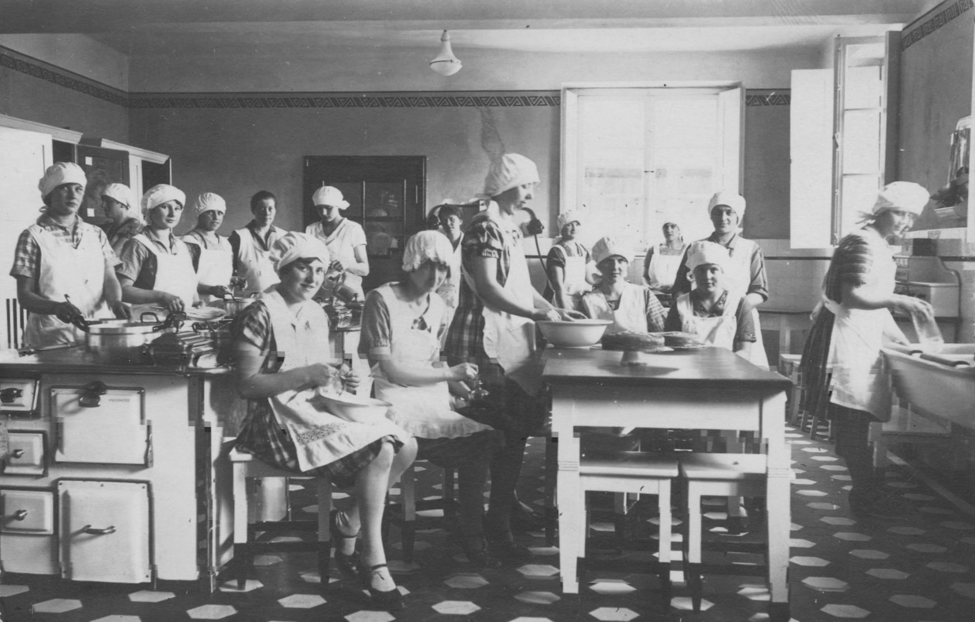 Schwarzweiß-Foto von jungen Frauen mit Haube und Schürze in einer Küche.
